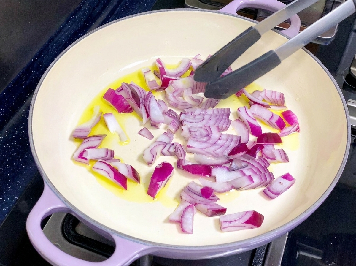 A light-colored pan containing olive oil and chopped red onions being stirred with tongs.