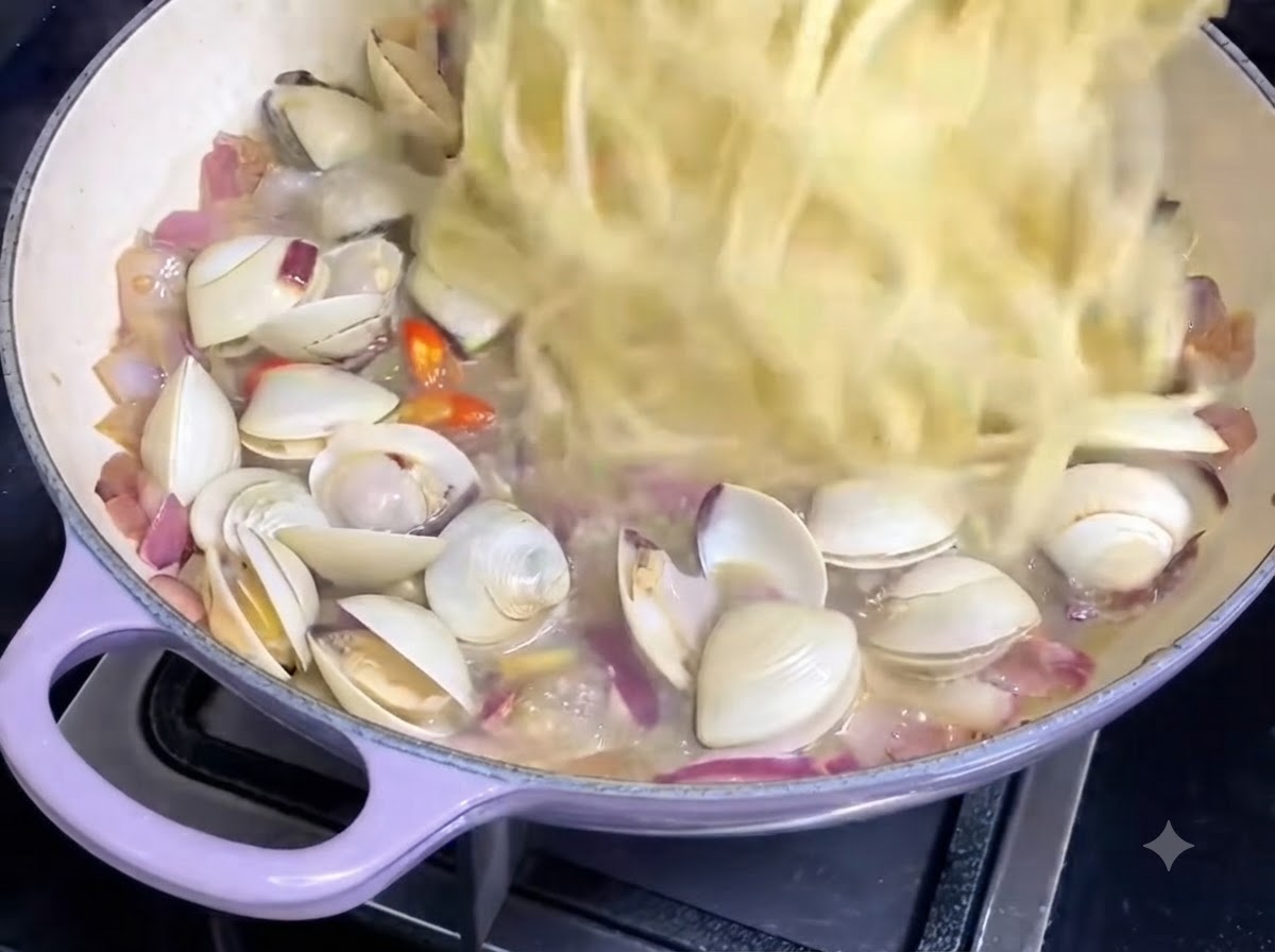Cooked spaghetti strands being tossed into a pan containing clam sauce and onions.