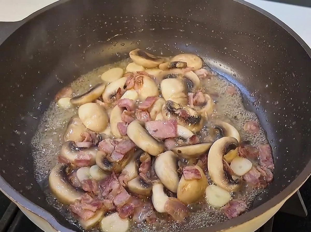 Cooked spaghetti strands being tossed into a large skillet with sautéed mushroom slices and bacon pieces.