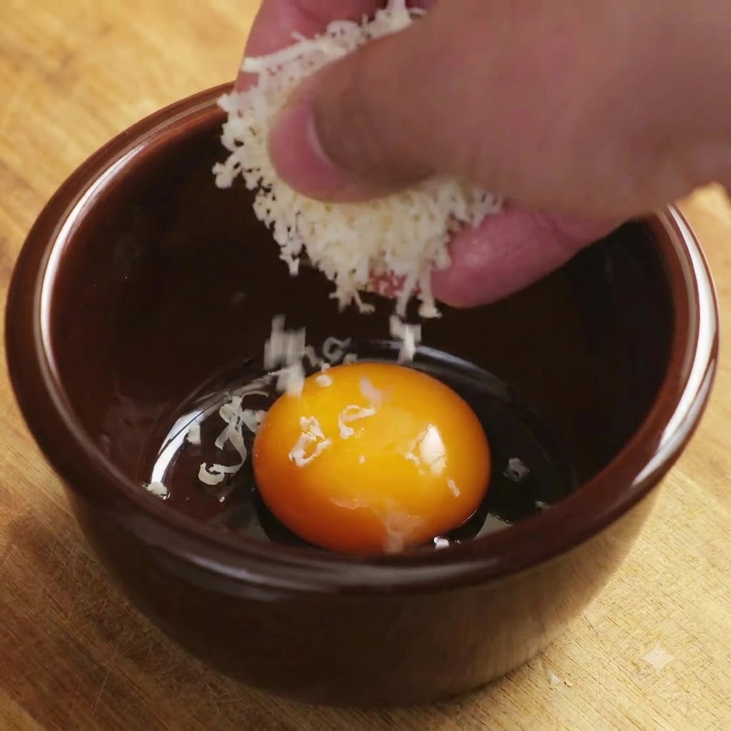 Grated Parmesan cheese falling onto a bright egg yolk inside a brown ceramic bowl.