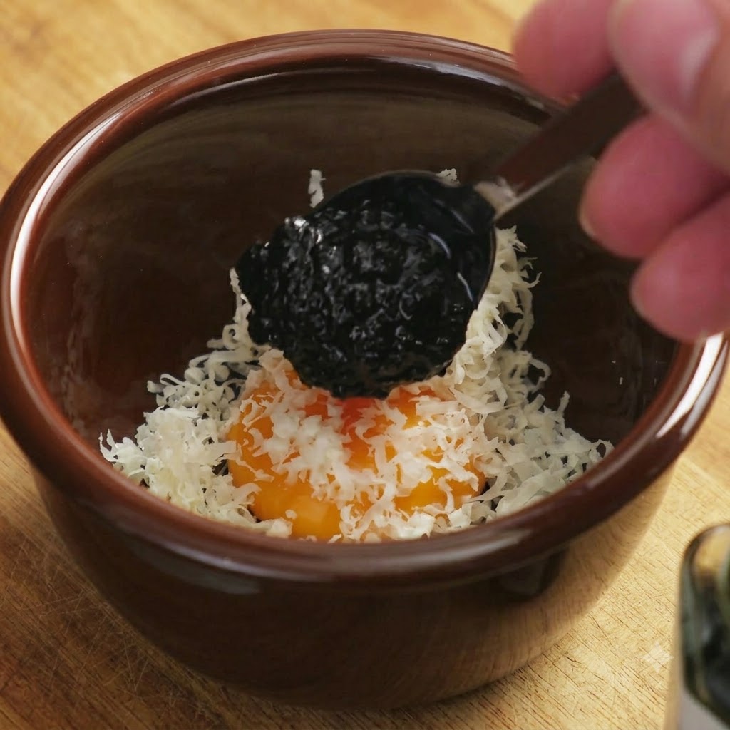 A dark spoonful of black truffle sauce being dropped onto the egg yolk and cheese mixture.