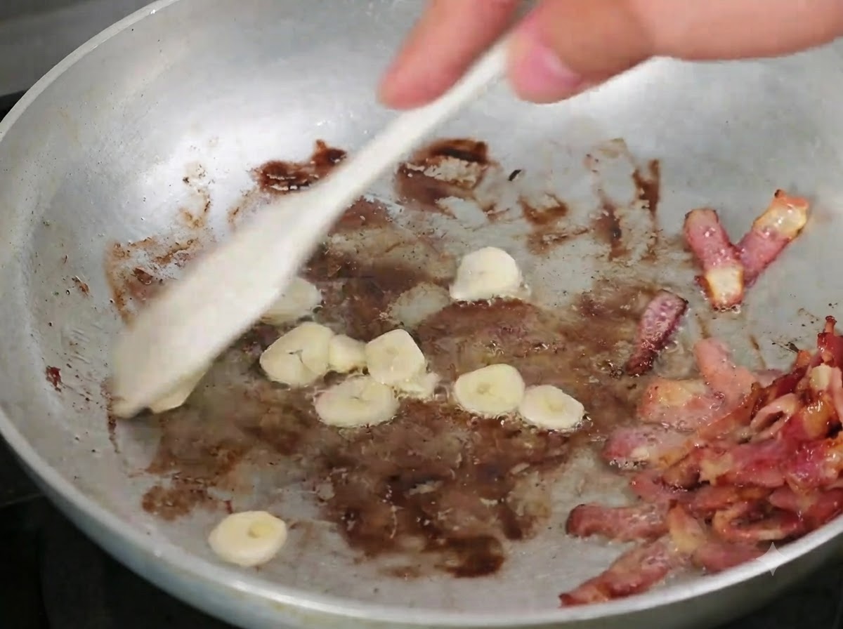 Sliced garlic being mixed into the pan with cooked bacon using a wooden spoon.