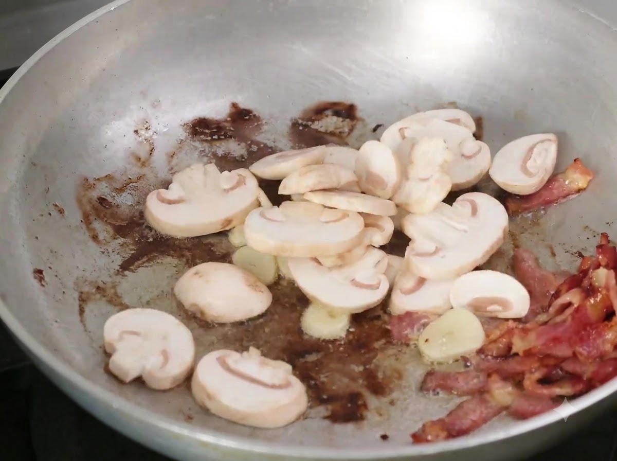 Sliced mushrooms being added to the frying pan with bacon and garlic.