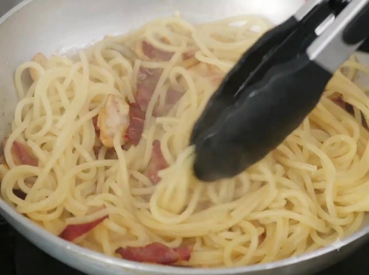 Butter and pasta water being mixed with spaghetti in a frying pan using tongs.
