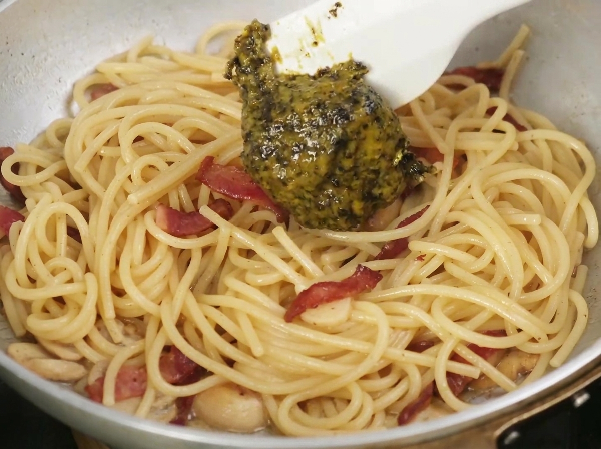 Black truffle sauce being spooned into a pan of cooked pasta and bacon.