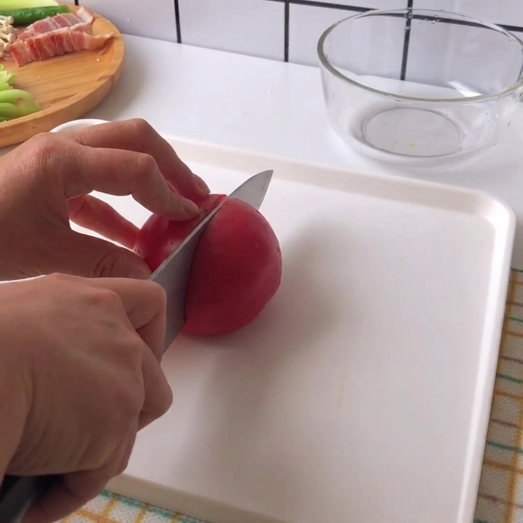 Hands holding a knife and slicing a fresh red tomato into chunks on a white cutting board.