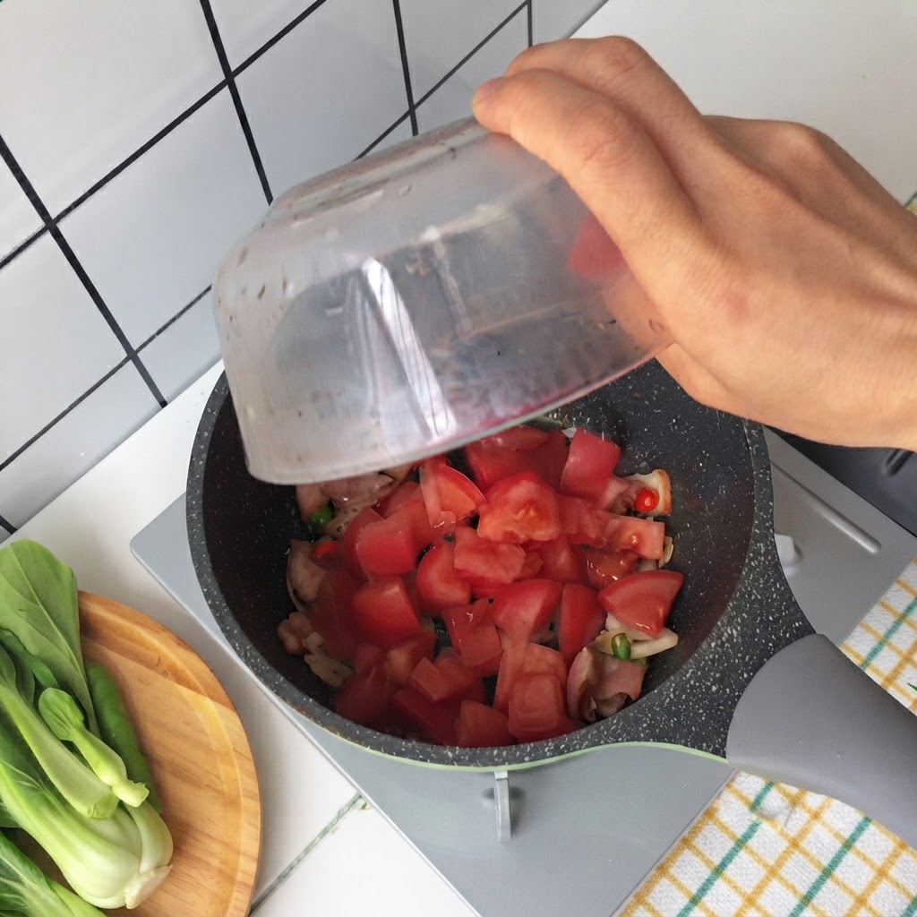 Hand pouring chopped tomatoes from a glass bowl into a pan containing fried bacon and green onions.