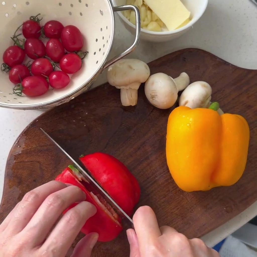 Red bell pepper being sliced on a wooden cutting board next to a yellow bell pepper and button mushrooms.