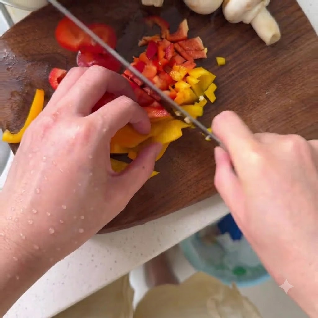 Fresh button mushrooms being sliced on a wooden cutting board next to diced colorful bell peppers.