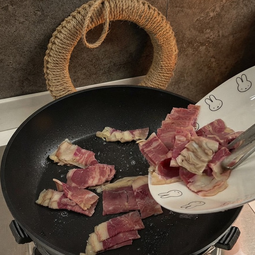 Sliced bacon pieces being added to and stir-fried in a black pan.