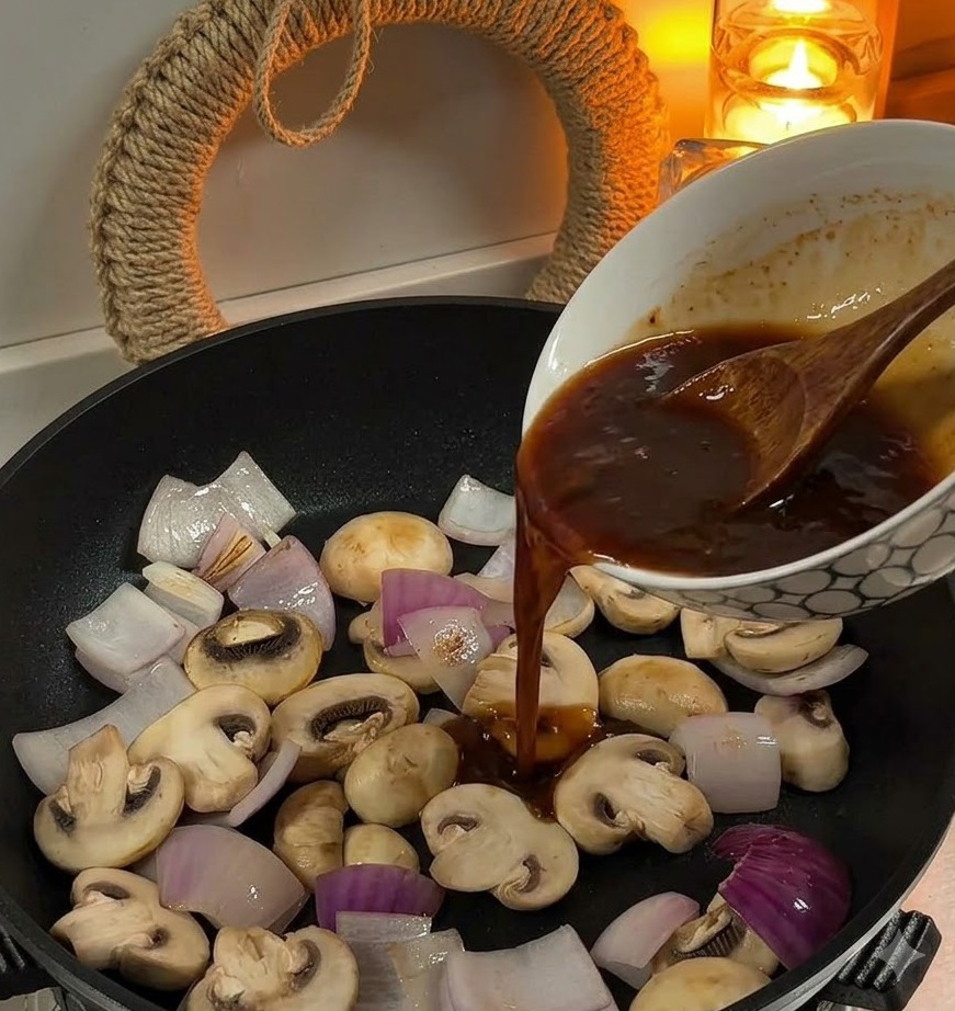Pouring brown sauce from a bowl into a pan containing sliced onions and white mushrooms.