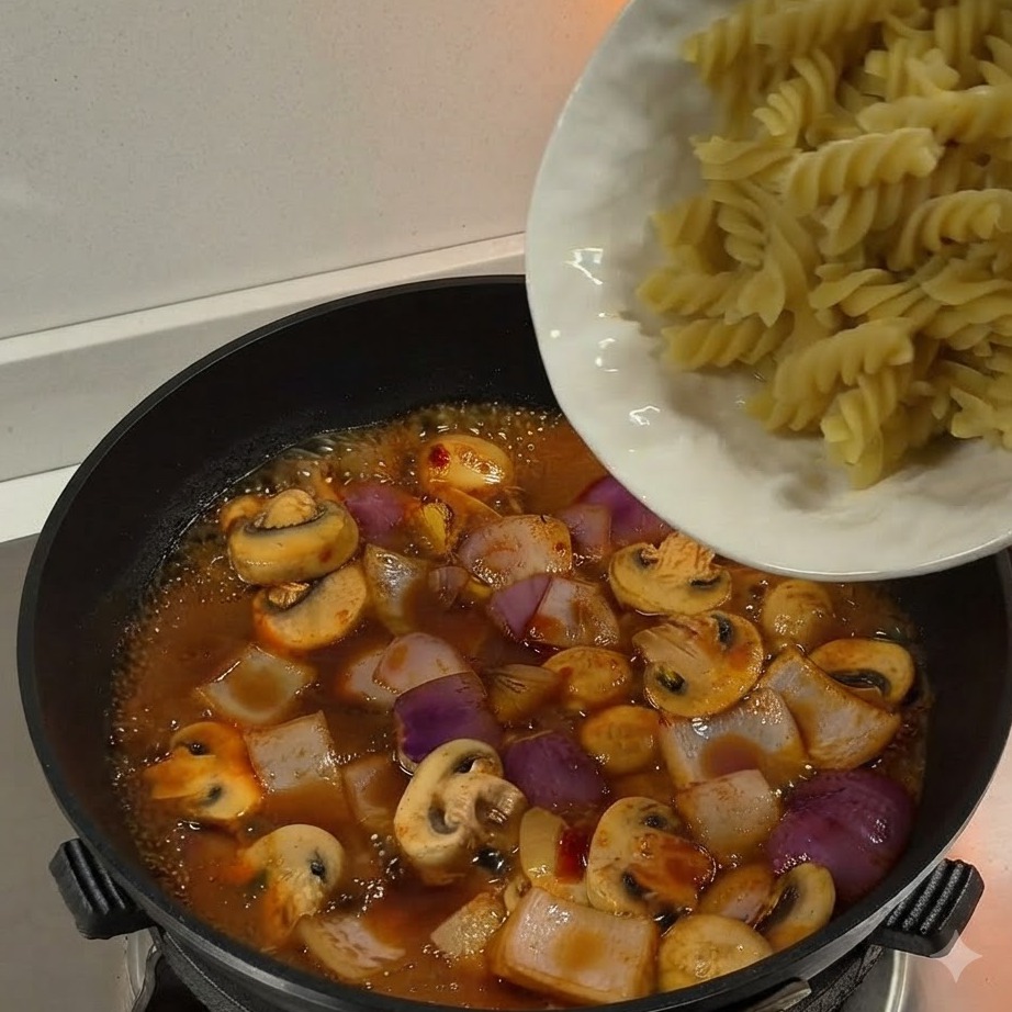 Boiled fusilli pasta being poured from a white plate into a pan filled with simmering sauce and vegetables.