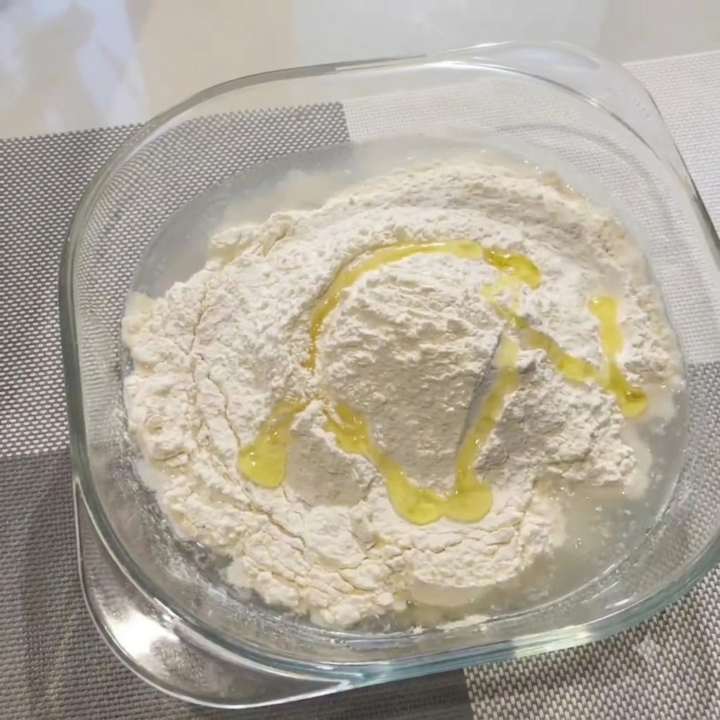 Olive oil drizzled over flour and water in a glass bowl.