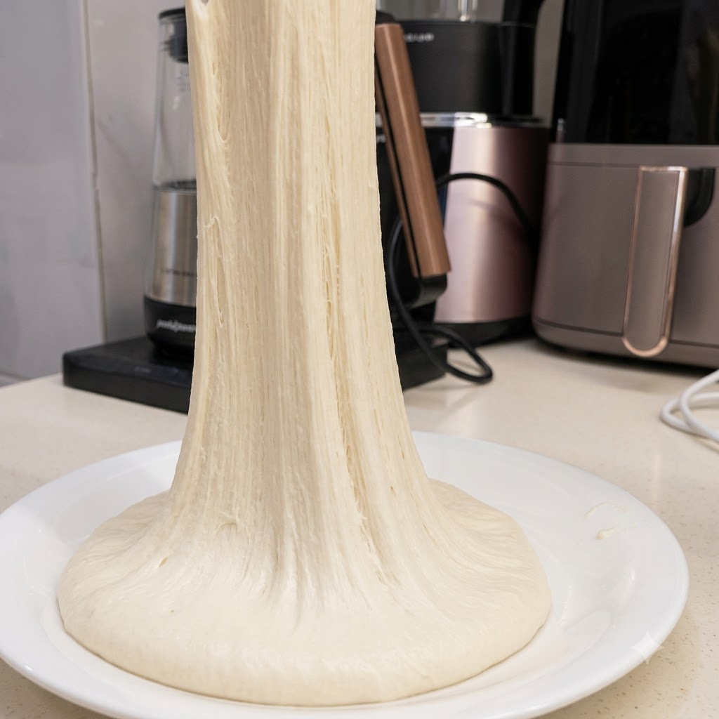 A long, continuous strand of highly elastic pizza dough being pulled upwards from a white plate.