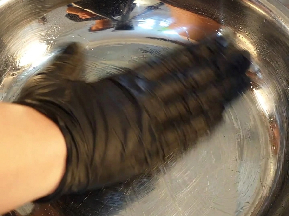 A hand wearing a black glove uses a paper towel to coat the inside of a shiny stainless steel bowl with oil.