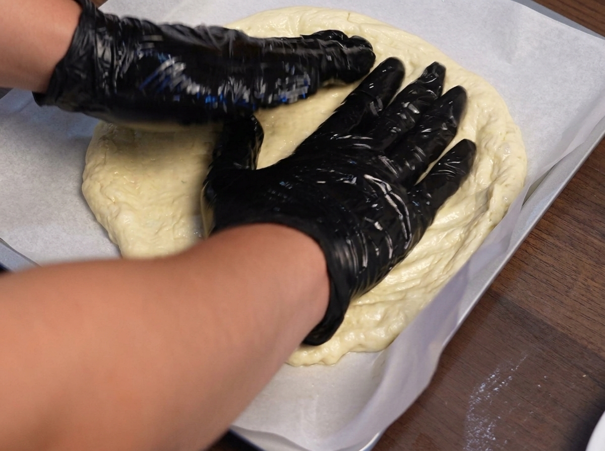 Gloved hands shaping pizza dough outward on a parchment-lined baking sheet.