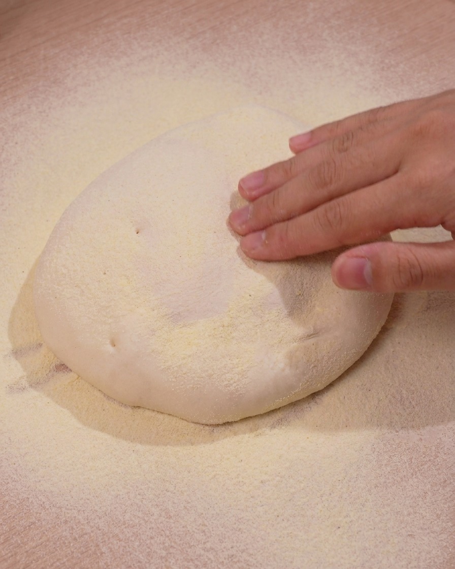 Hand pressing down gently onto a mound of pizza dough resting on yellow cornmeal.
