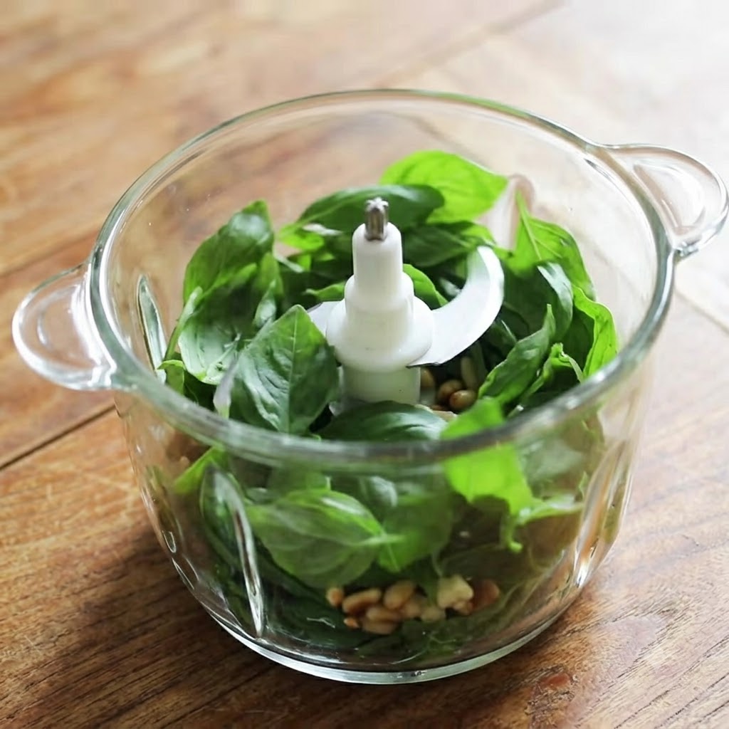 A glass blender container filled with fresh basil leaves, pine nuts, garlic, and cheese ready for processing.