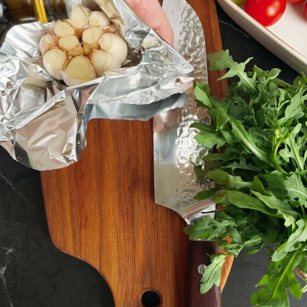 Olive oil being drizzled onto an unwrapped garlic bulb resting on aluminum foil.