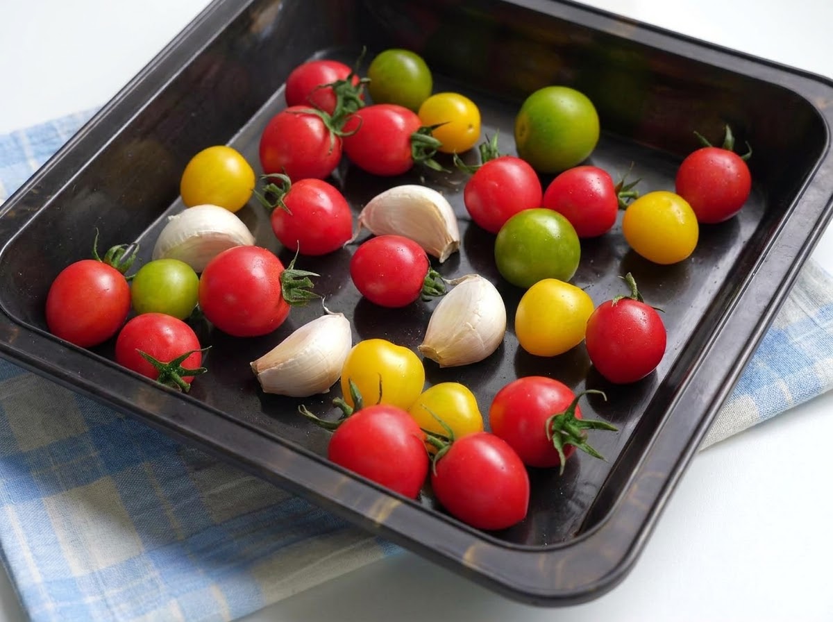 A baking tray filled with colorful cherry tomatoes and garlic cloves coated in olive oil and seasonings.