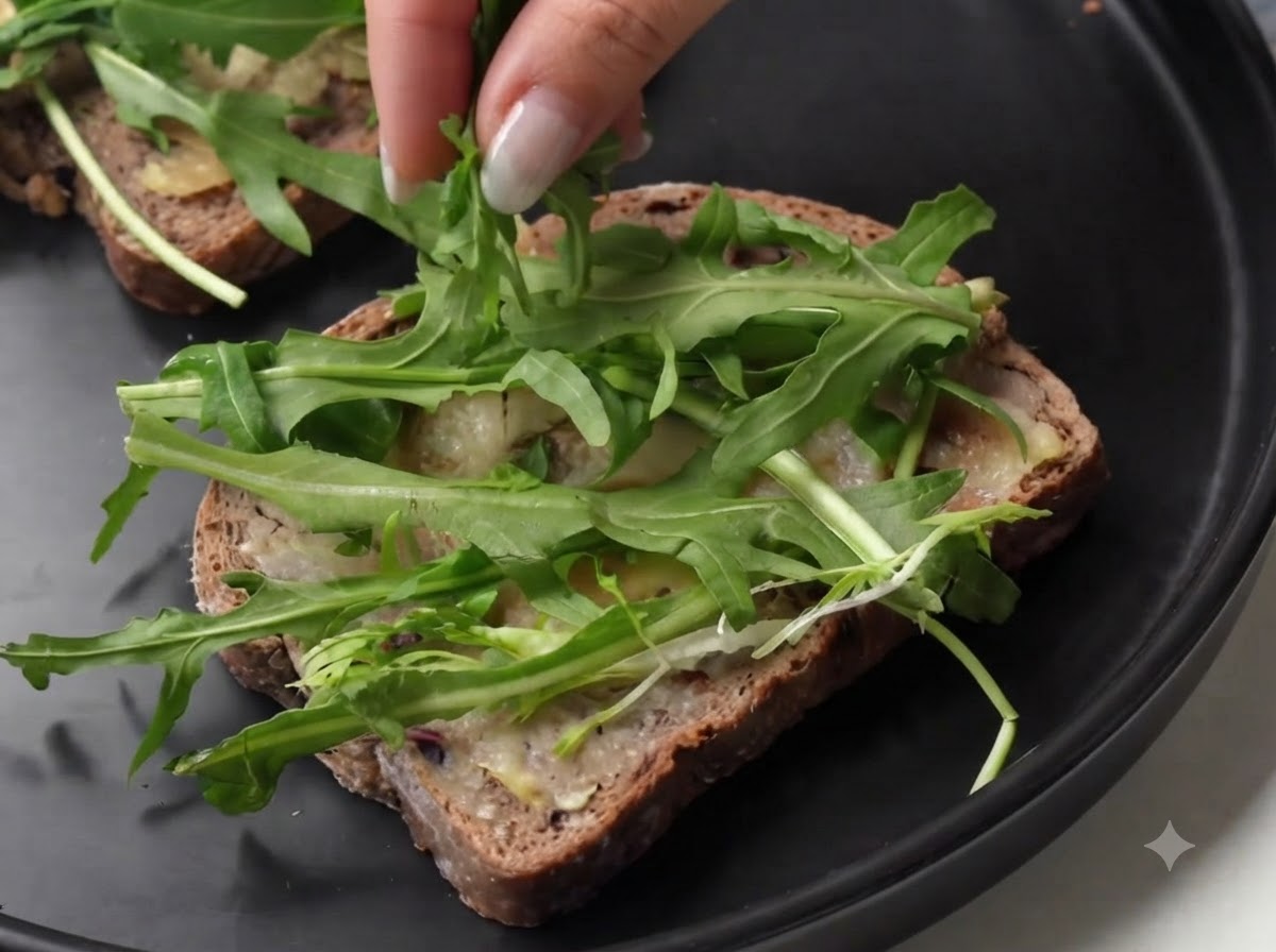 A person layering slices of cured ham over fresh green arugula on a slice of bread.
