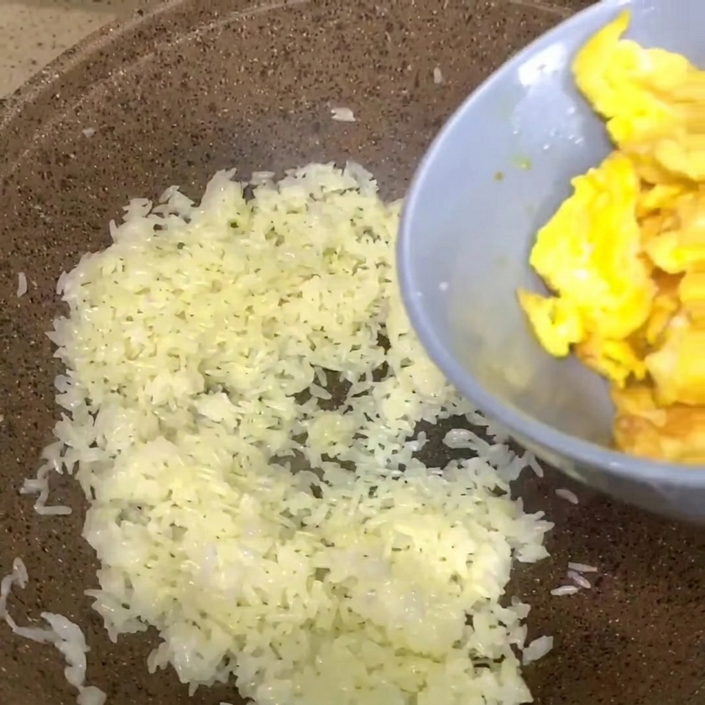 Scrambled eggs from a grey bowl being poured into a pan of white cooked rice.
