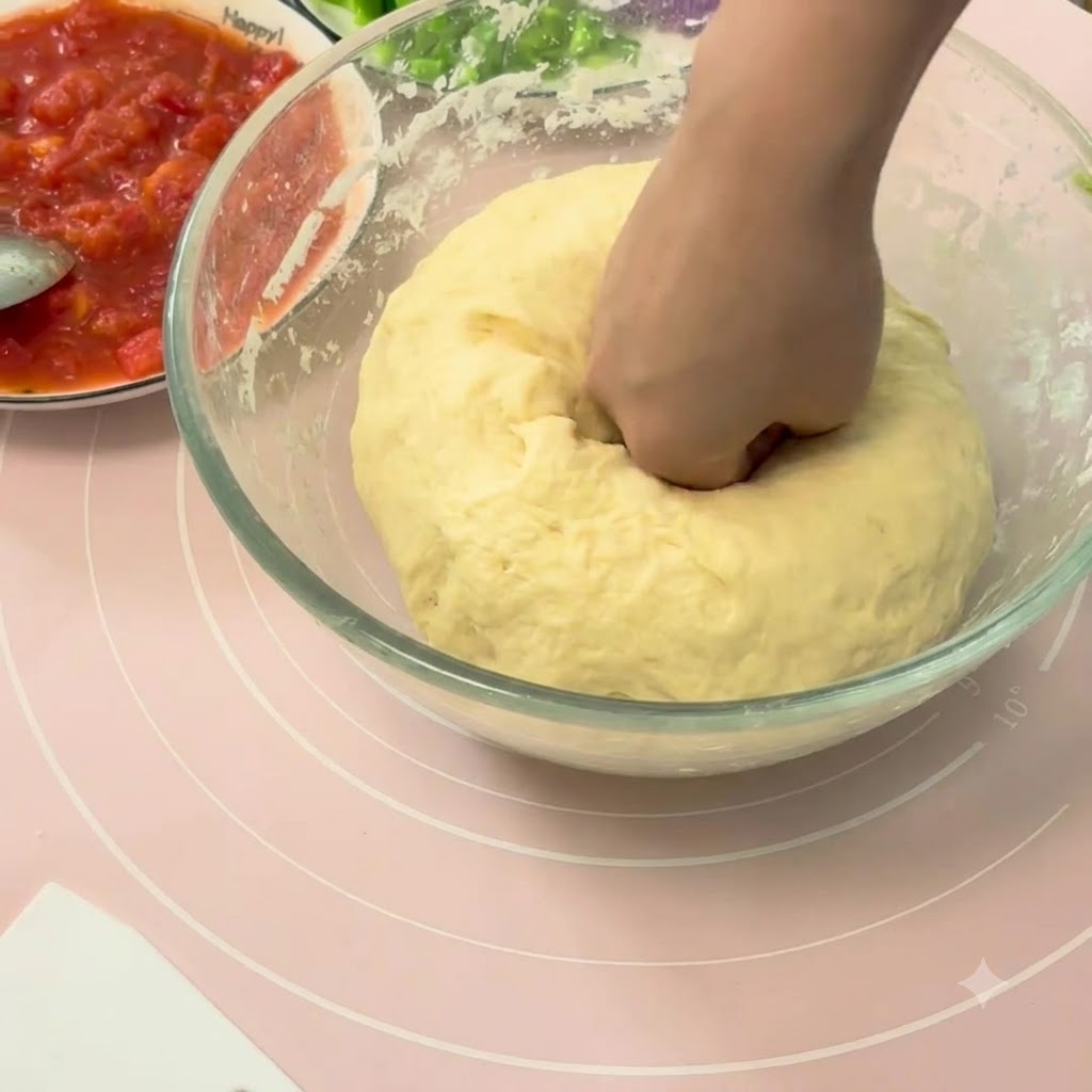 A hand punching down a large ball of risen pizza dough inside a glass bowl.