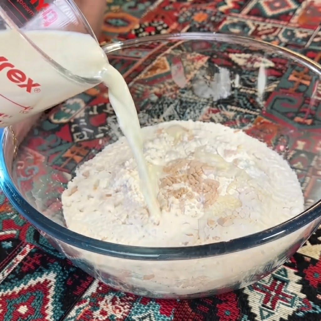 Milk being poured from a clear glass measuring cup into a bowl of flour and yeast.