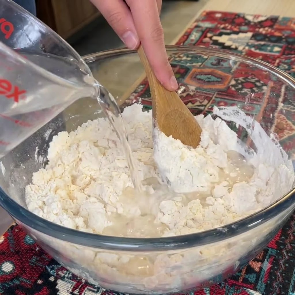 Water being poured from a measuring cup into a glass bowl of flour while a wooden spoon stirs.