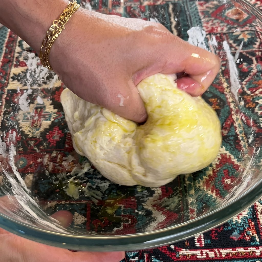 A hand firmly kneading and folding dough mixed with olive oil inside a glass bowl.