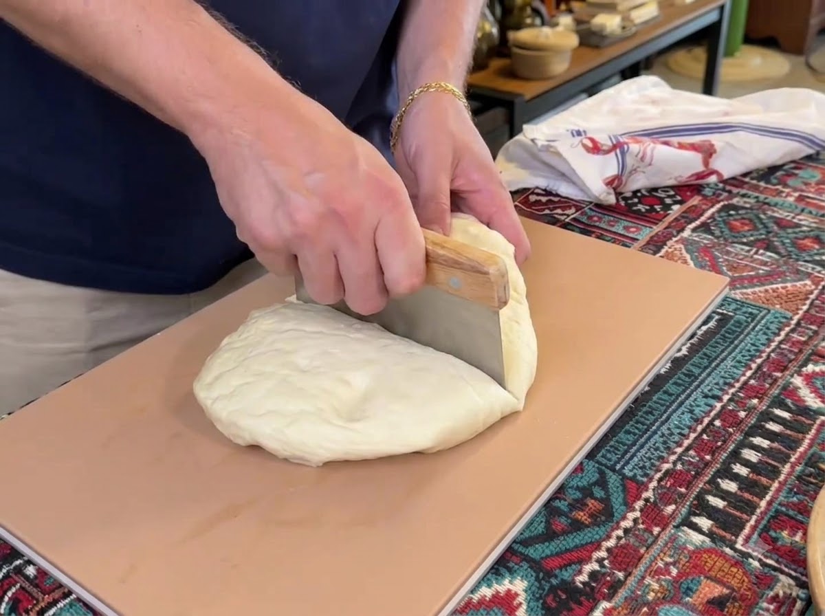 A dough scraper cutting a large mound of white pizza dough into four separate pieces on a cutting board.