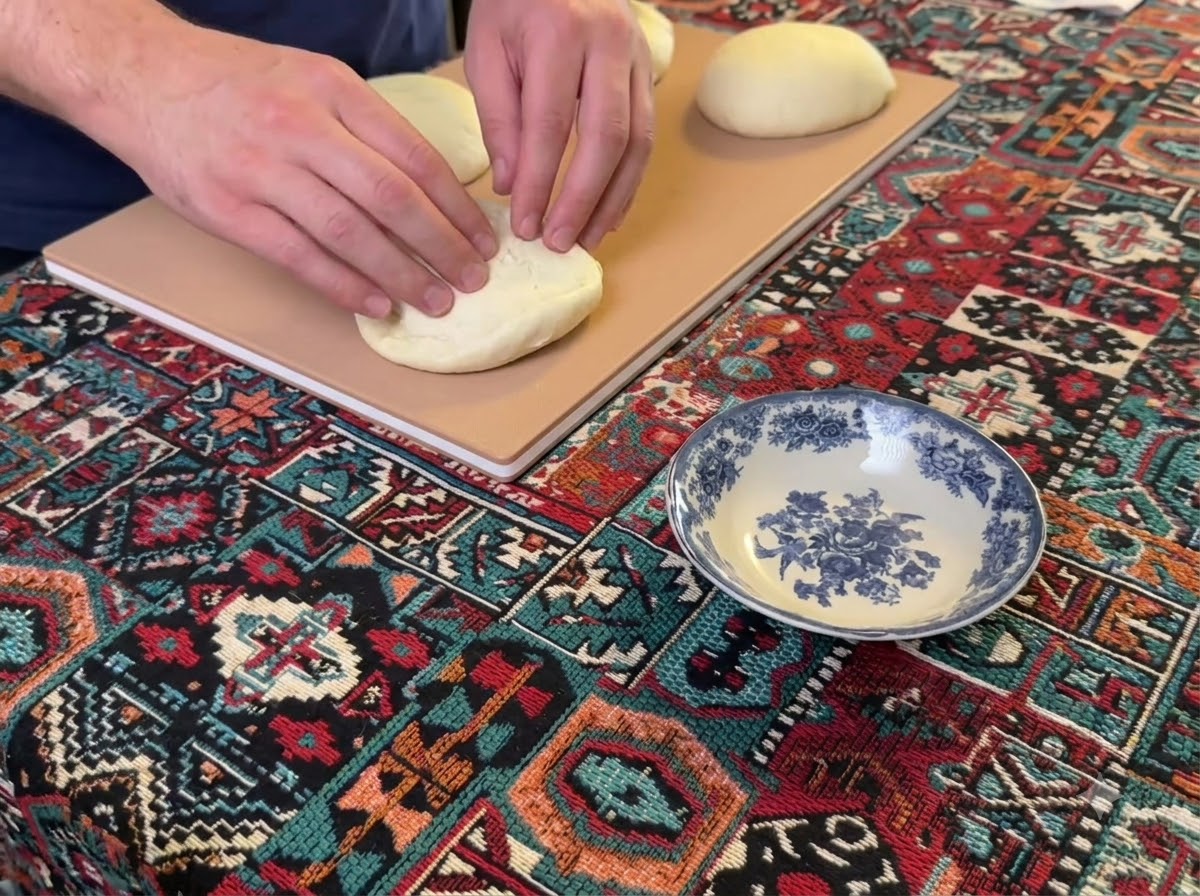 Hands pressing down and flattening a round ball of pizza dough on a wooden board.