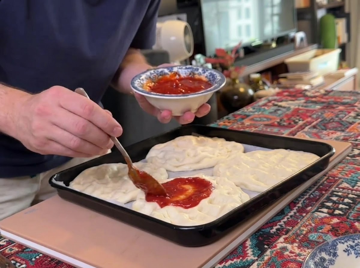 A spoon spreading red tomato sauce onto multiple flattened pizza dough bases laid out on a black baking tray.