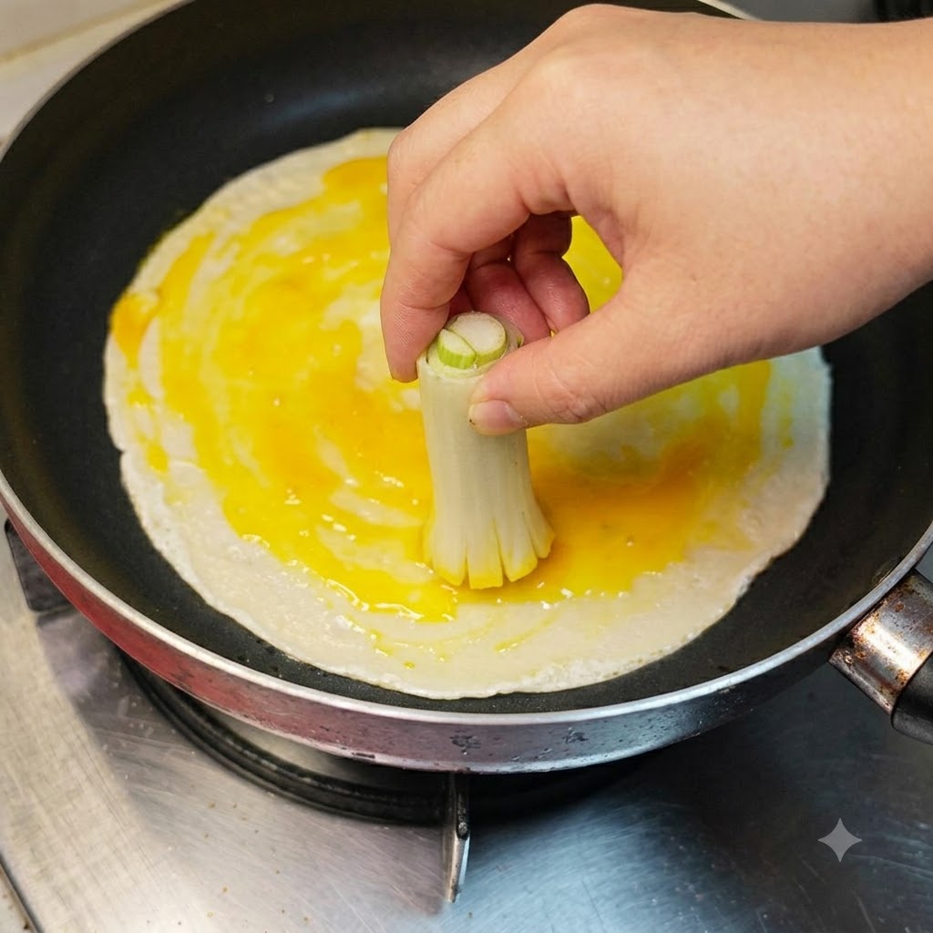 A hand using a tied bunch of scallion stalks to brush cracked egg white and yolk across a pancake.