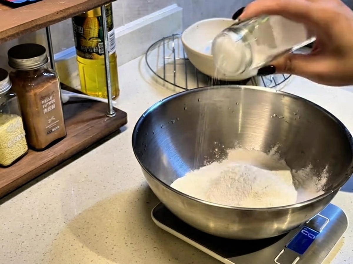 A hand shaking a glass jar to sprinkle salt over a mound of white flour inside a large metal mixing bowl.