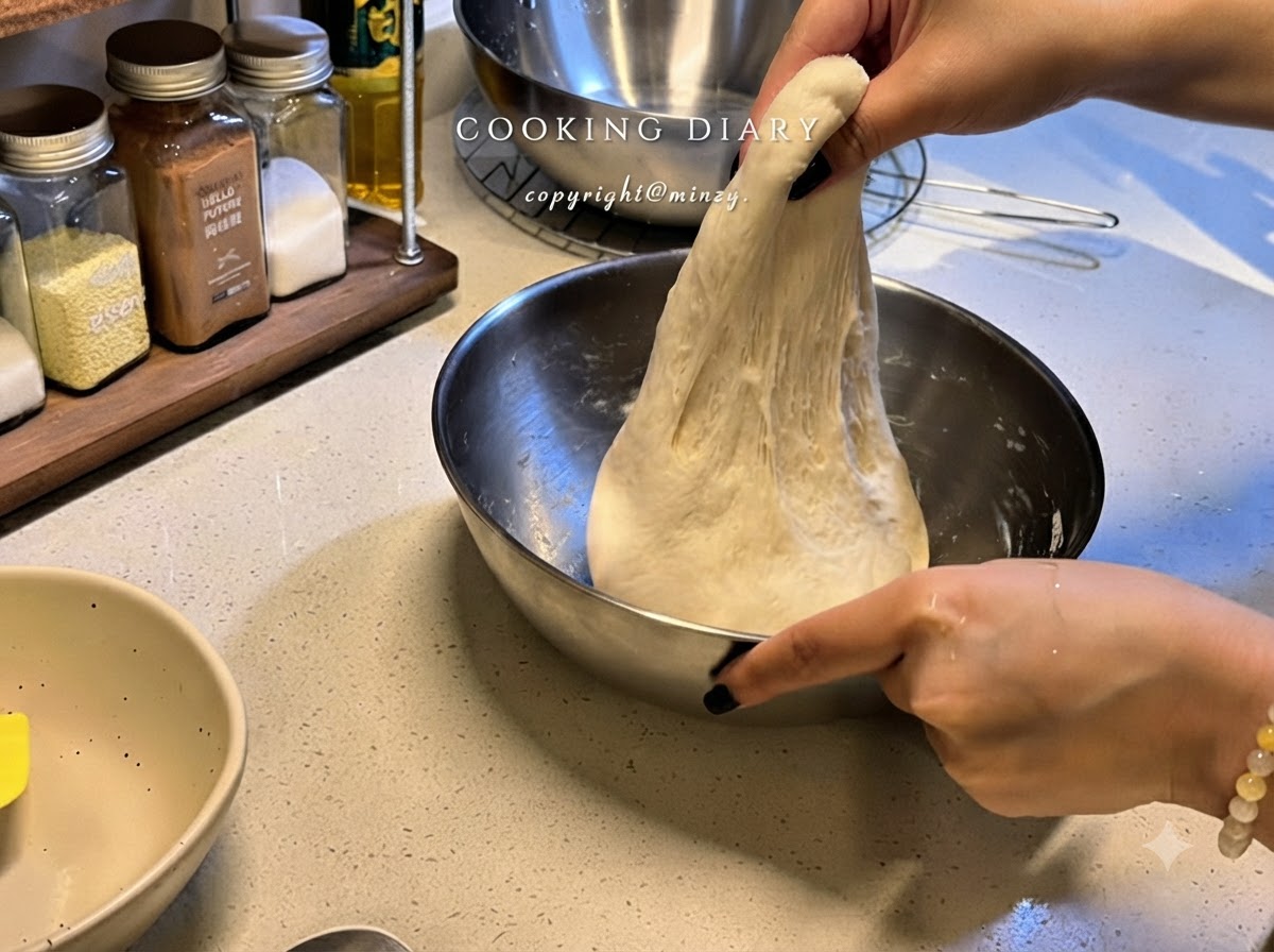 A pair of hands lifting a portion of elastic pizza dough upward inside a metal bowl to stretch it.