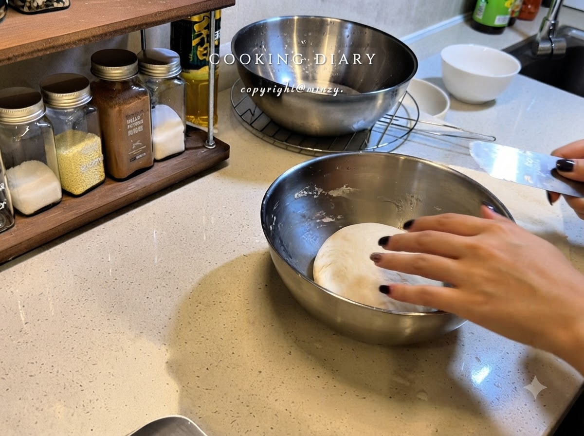 A hand smoothing plastic wrap over a stainless steel bowl containing a round ball of pizza dough.