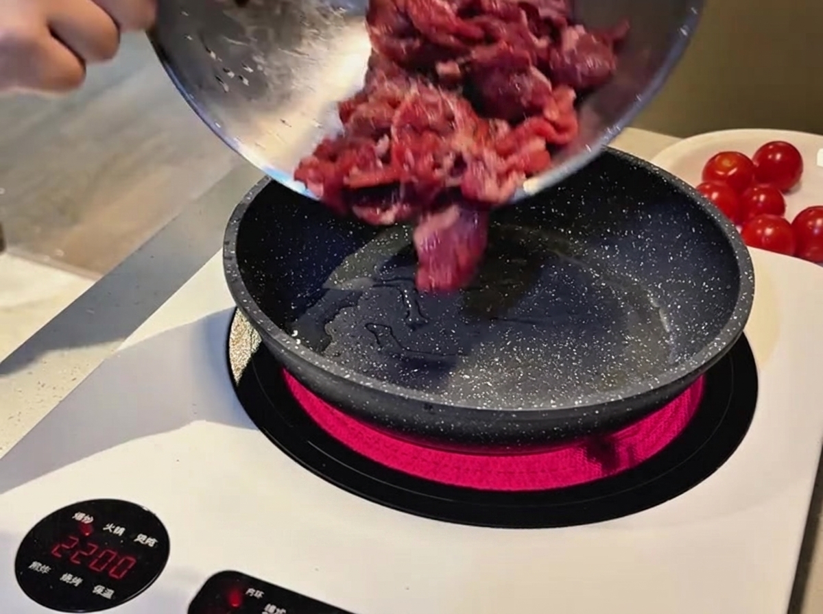 Raw, marinated sliced beef being poured from a metal bowl into a hot speckled frying pan on an induction cooktop.