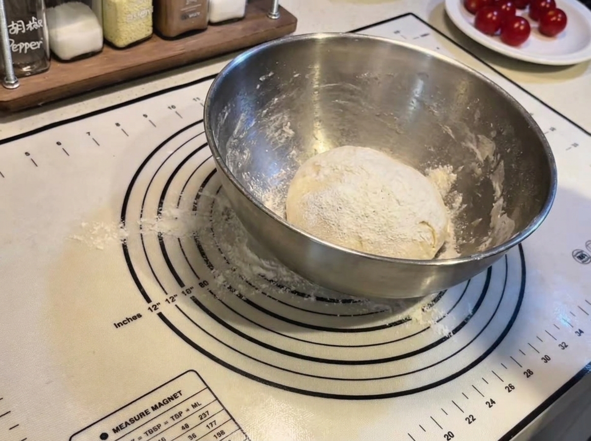 A round ball of pizza dough dusted with flour inside a steel bowl on a silicone baking mat.