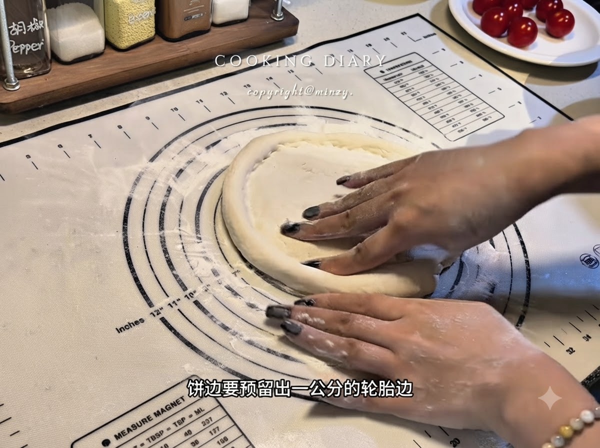 Hands shaping the edges of a flat pizza dough base on a silicone mat to form a raised rim.