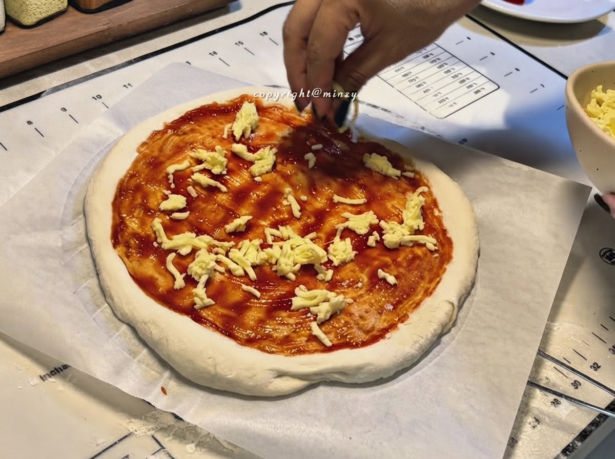 Hands sprinkling shredded cheese onto a pizza base coated with red pizza sauce.