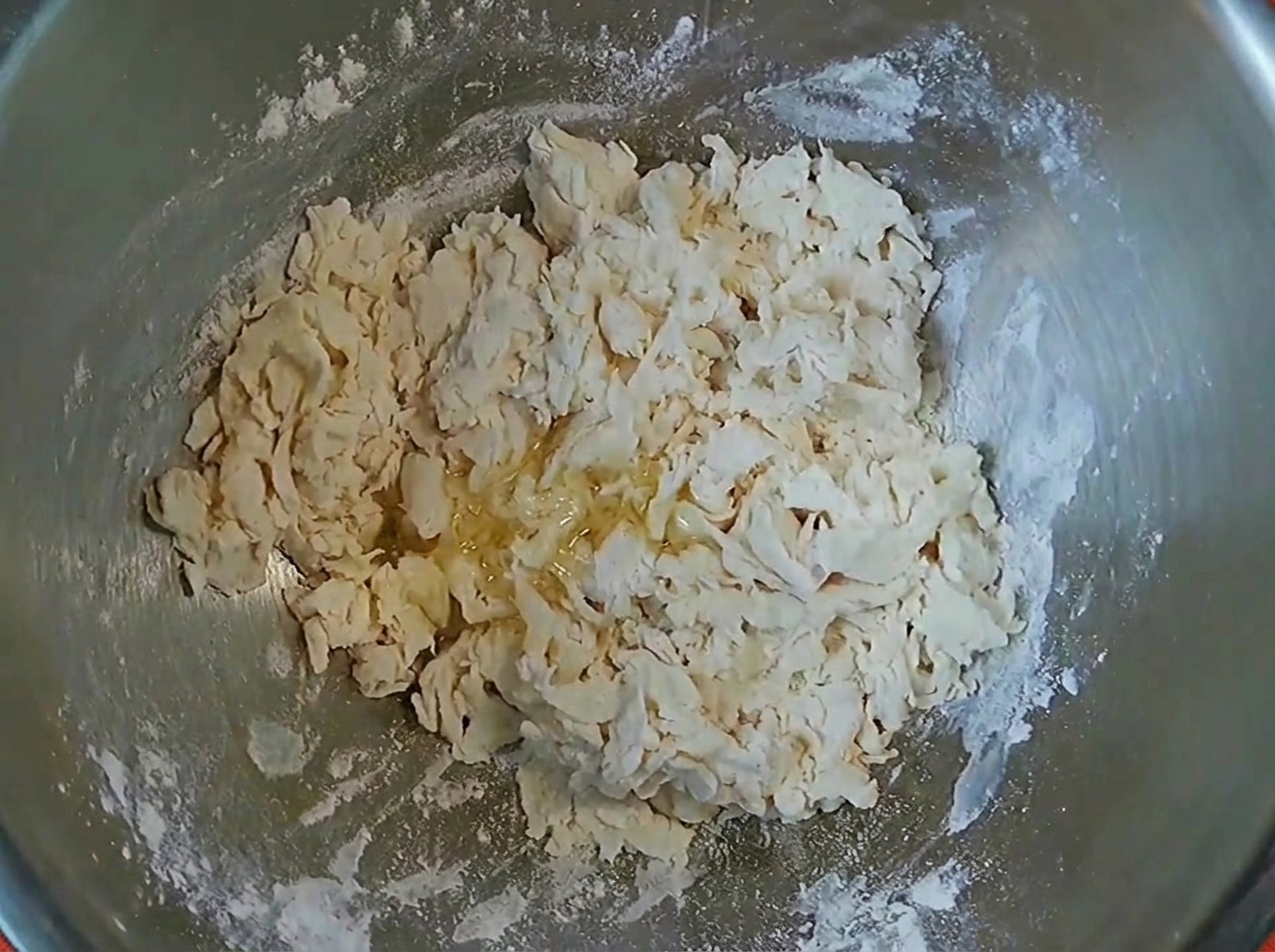 Dough flakes with a drizzle of cooking oil inside a metal mixing bowl.