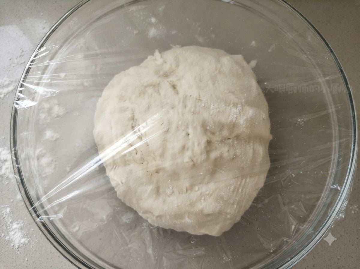 A smooth dough ball inside a clear glass bowl covered tightly with transparent plastic wrap.