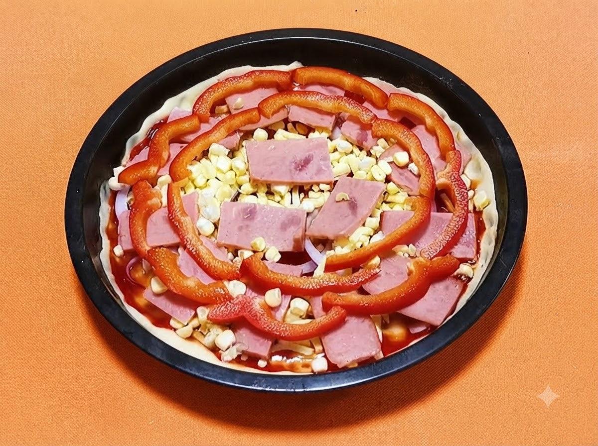 Pizza crust in a black pan topped with sauce, ham, yellow corn kernels, and red bell pepper slices.