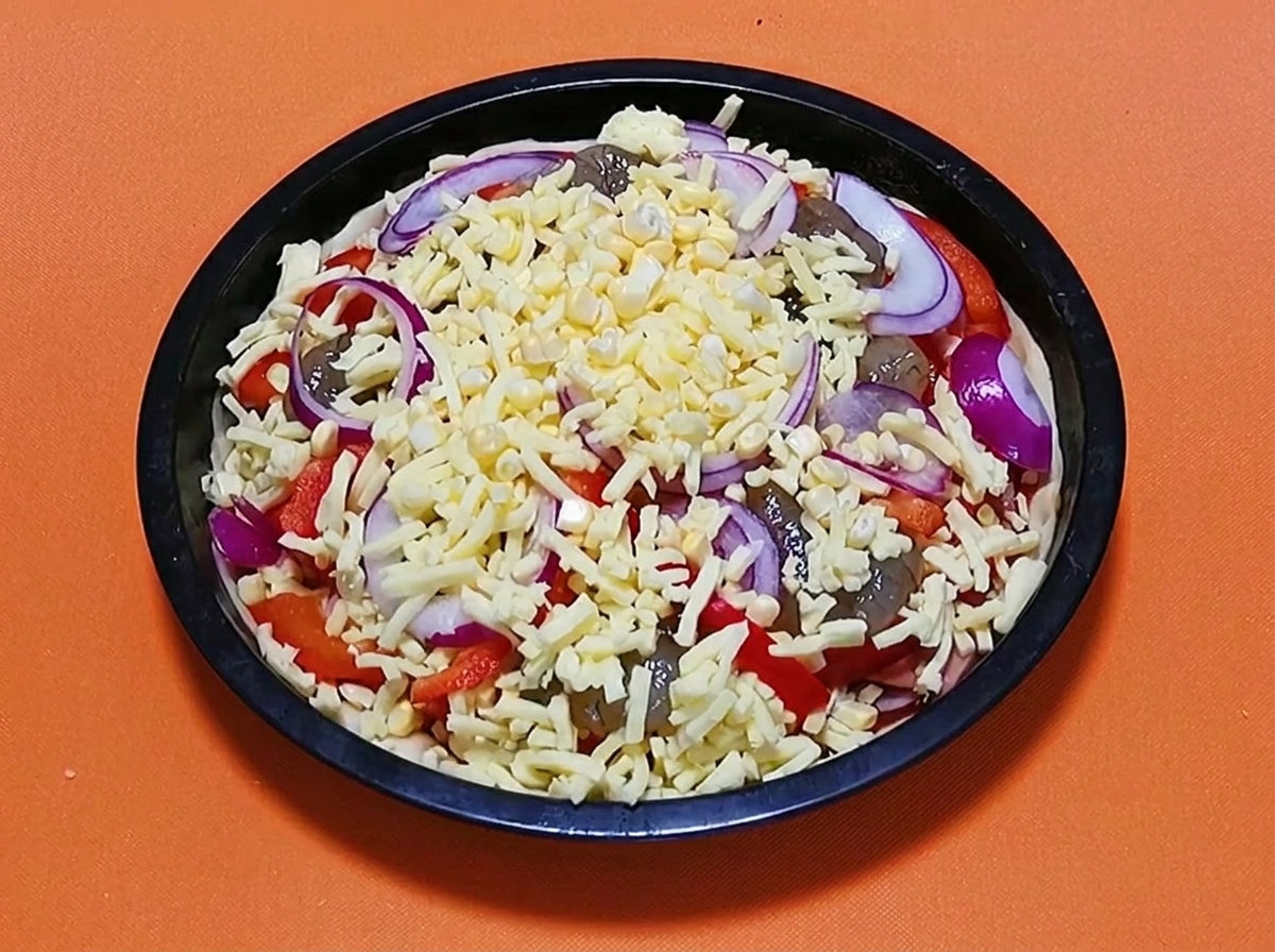 Fully assembled raw seafood pizza topped with a final layer of shredded cheese and red onion slices, ready to be baked.
