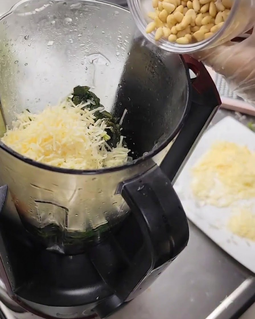 Pine nuts being poured from a small cup into the blender filled with cheese and basil.