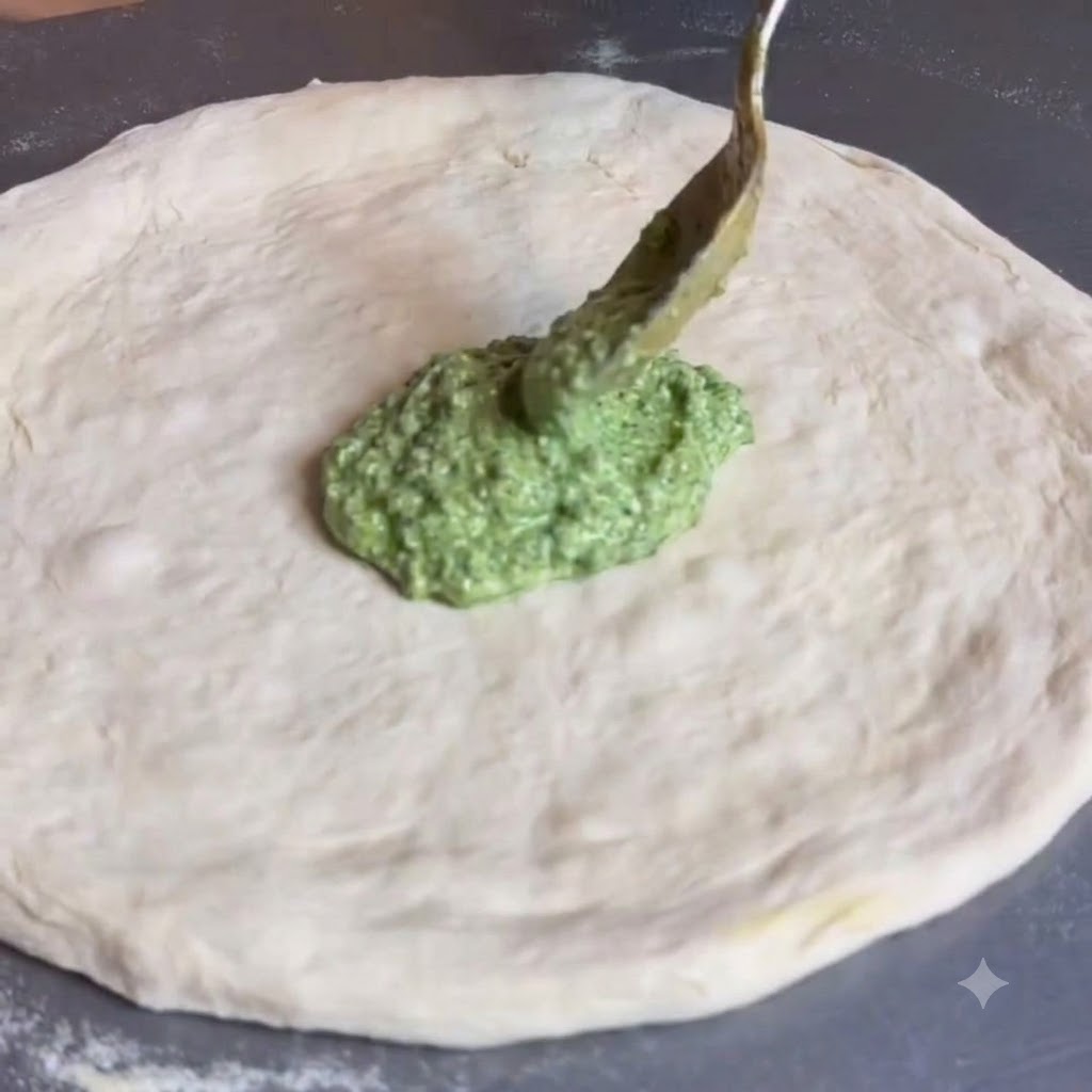 A spoon spreading green pesto sauce across the middle of a flat round pizza dough.
