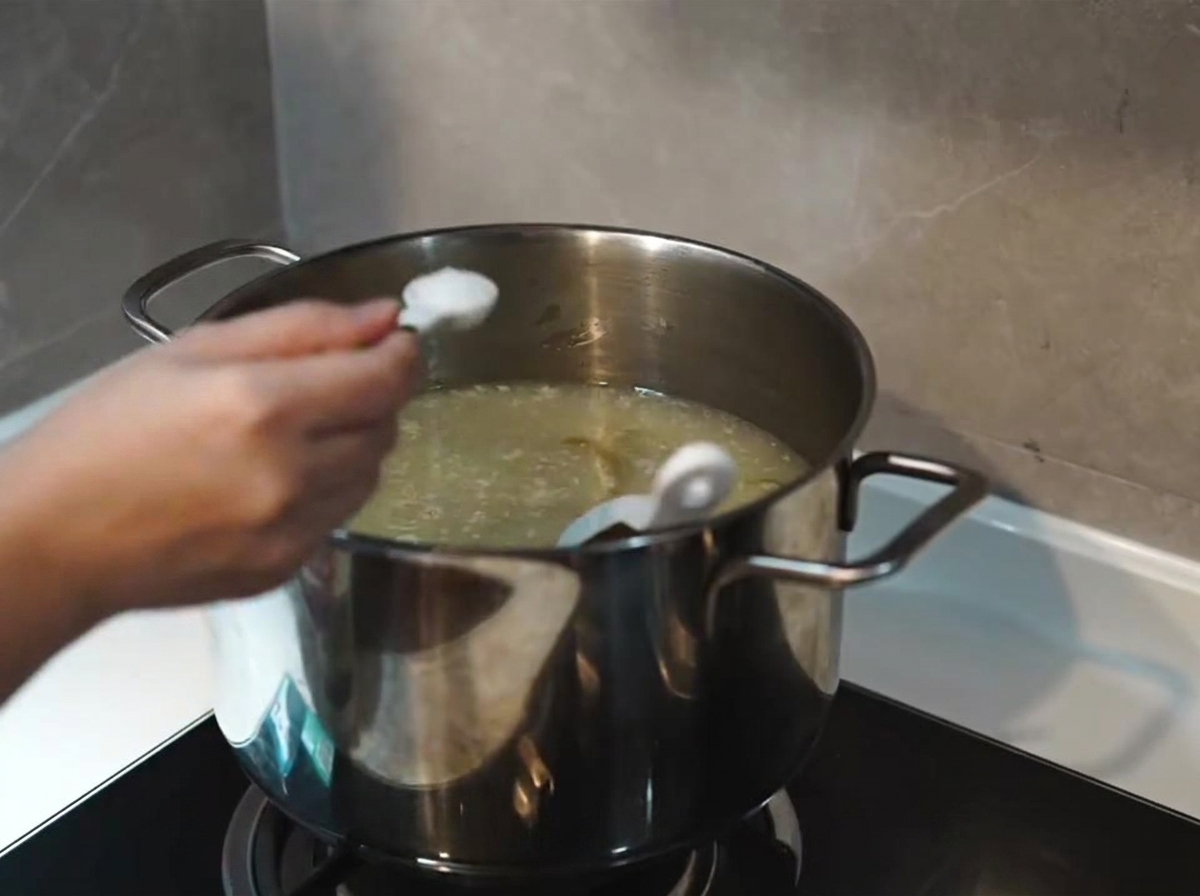 A spoonful of salt being added to a simmering, thickened milky-white soup base in a stainless steel pot.