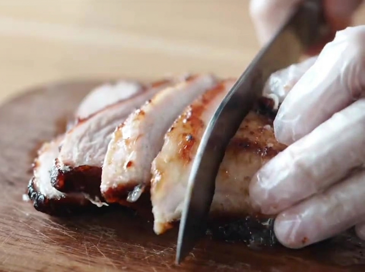 A piece of caramelized, honey-glazed smoked chashu pork being sliced into thick pieces by someone wearing gloves on a wooden cutting board.