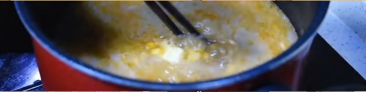Chopsticks stirring a vigorously boiling orange miso broth with corn and a pat of butter in a dark pot.